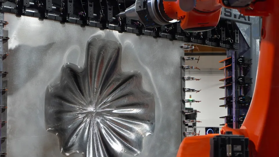 An orange KUKA robot arm holds a forming tool above a clamped aluminum sheet bearing a cross-shaped radial relief at Machina Labs.