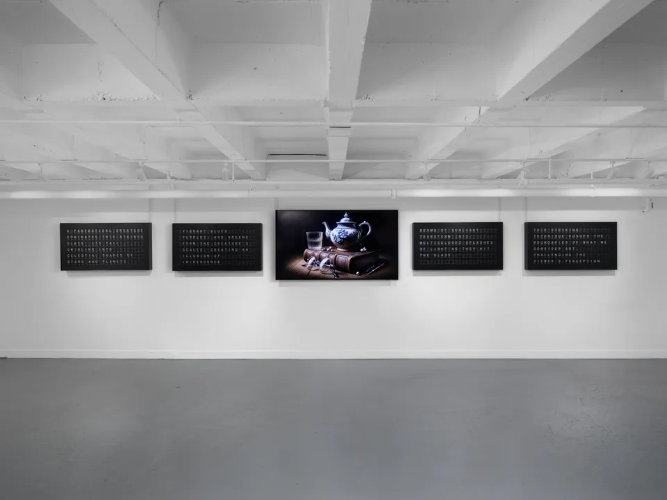 Four black split-flap text displays flank a monitor showing a teapot, glass, and books beneath white ceiling beams.