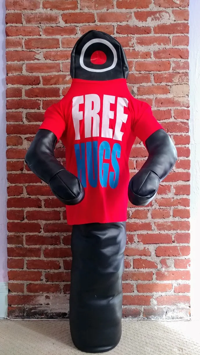 A full-length grappling dummy in a red Free Hugs T-shirt standing against a brick wall