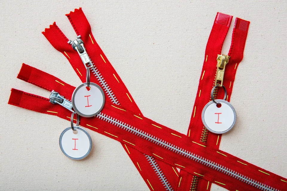 Close view of three red zipper pulls carrying round tags marked I, with yellow stitching visible on the zipper tape.