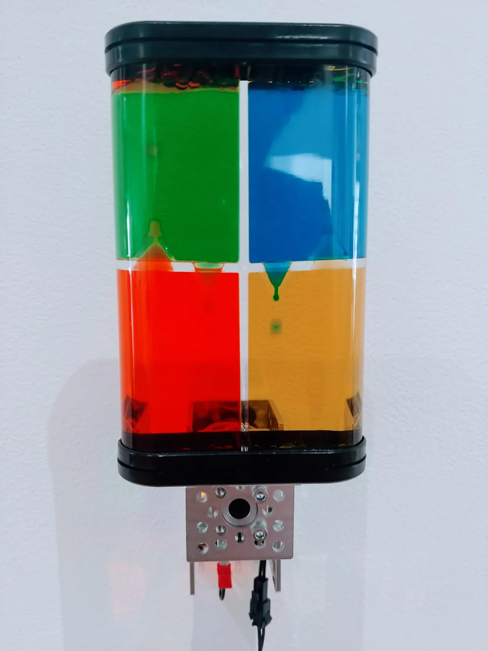 A transparent toy with dark end caps contains green, blue, red, and yellow liquid chambers above a small motor.
