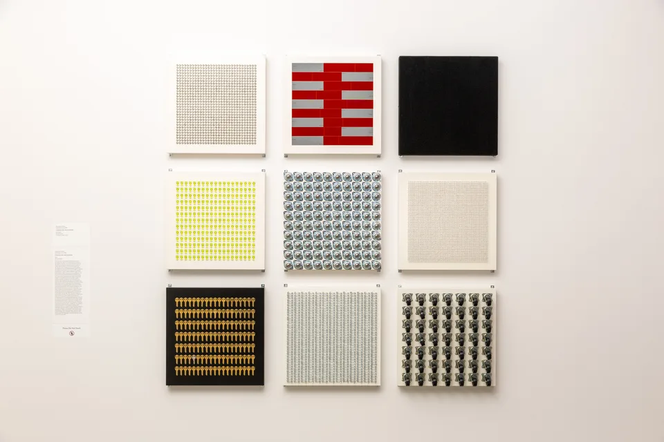 Nine square panels of repeated manufactured objects hang in a three-by-three grid on a white wall.
