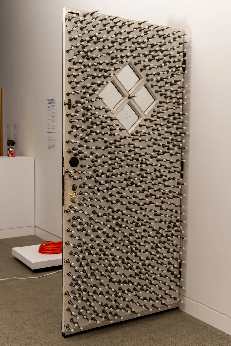 A white door covered with hundreds of projecting spring doorstops stands open in the gallery.
