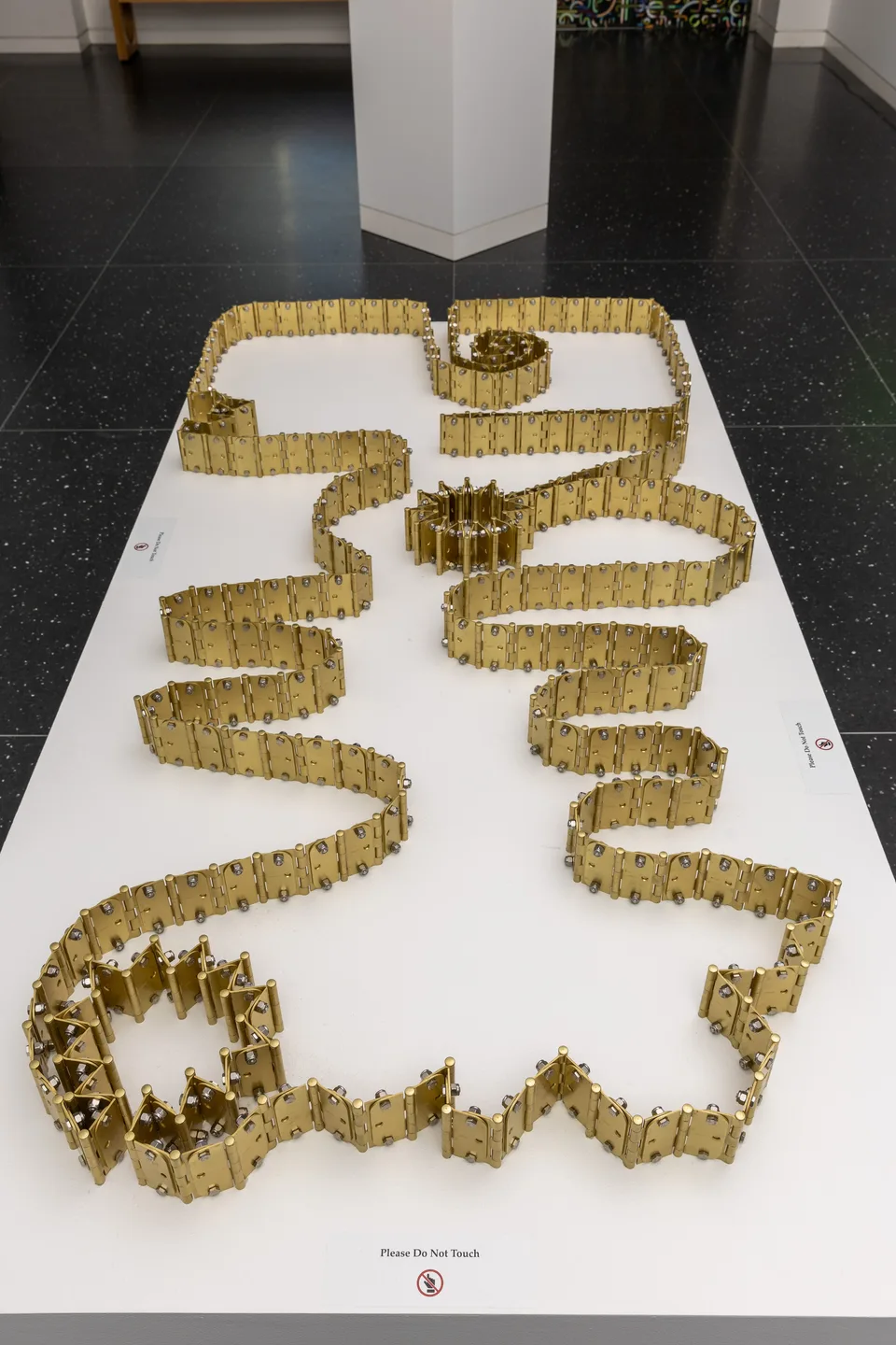Long connected chains of brass hinges arranged in winding paths on a white plinth.