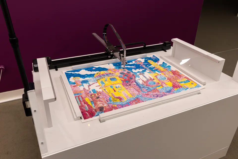 A pen plotting machine sits over a brightly colored drawing on a white table-like plinth.