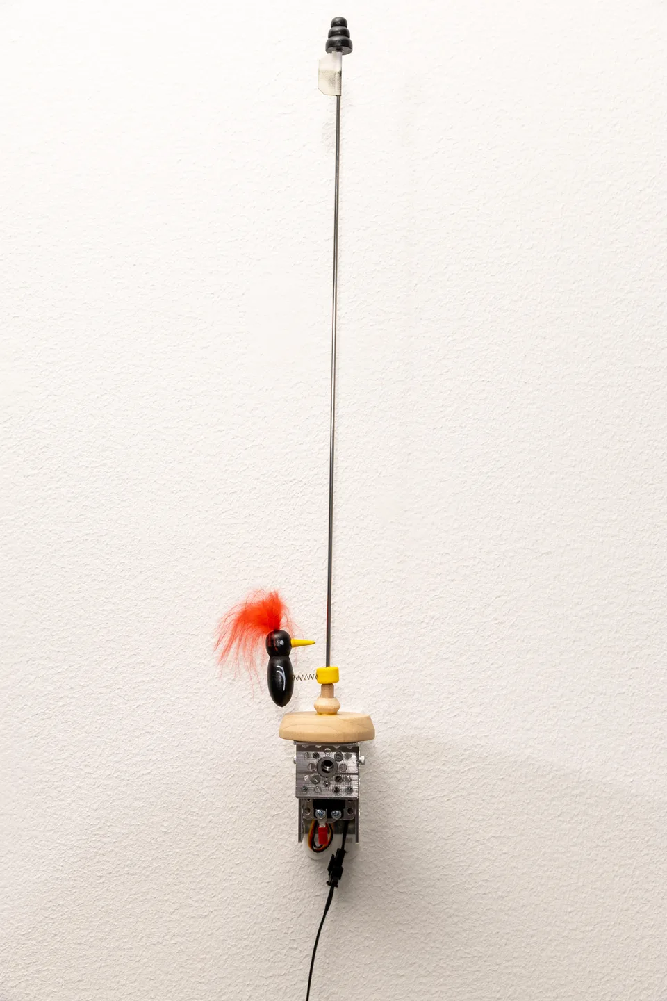 A small wooden bird with an orange tuft sits on a tall, slender rod above a wall-mounted motorized mechanism.