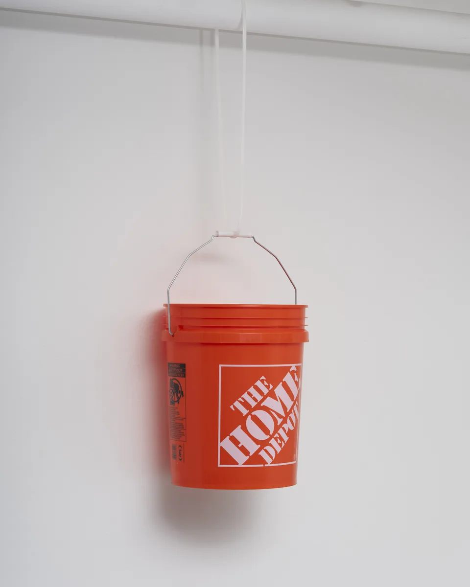 An orange Home Depot bucket hangs by its metal handle from a white cord against a white wall.