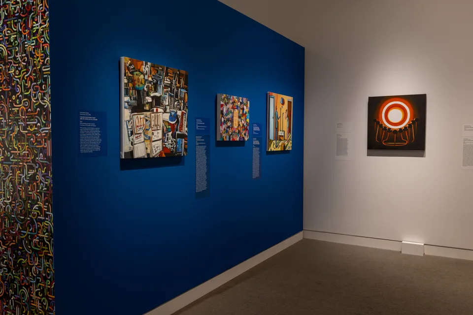 A Bunch of Items That Are on Display in a Store is the largest painting at left on the blue wall, shown with three other paintings.