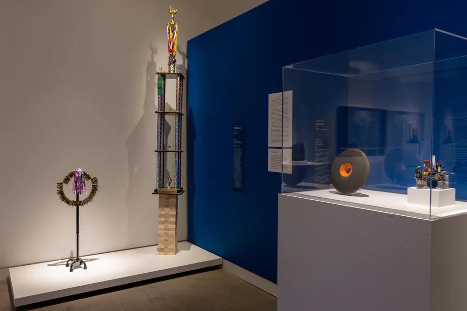 A tall trophy and a key-covered cane stand beside a vitrine containing a dark ceramic ring and a small assembly of electronic parts.