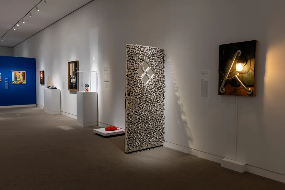 A freestanding door covered in spring stops stands beside a lit assemblage with metal rails; a coiled extension cord and a flip clock on a can are nearby.