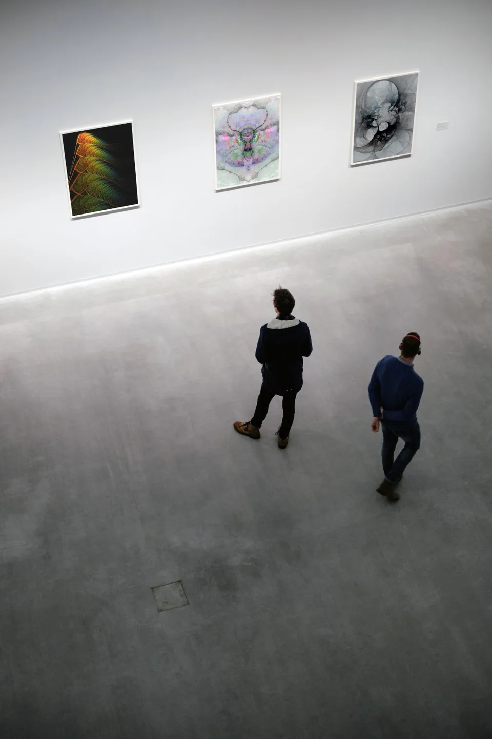 Three prints hang along a white gallery wall, with two visitors standing below.