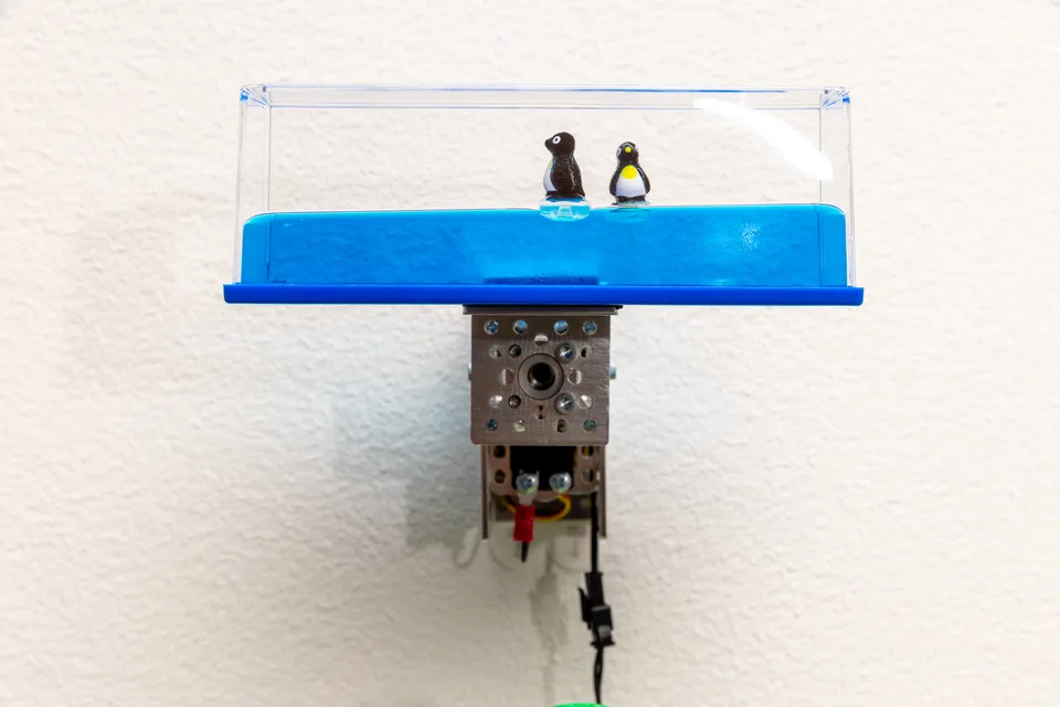 A transparent rectangular toy containing blue liquid and two floating penguins is mounted above a motorized mechanism.