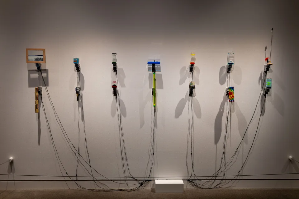 Seven columns of motorized toys, liquid timers, small displays, and trailing wires are arranged on the gallery wall.