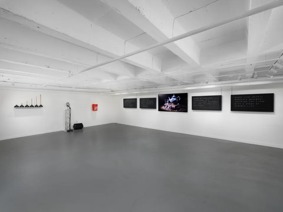 Wide view of the basement gallery with plungers, a metal dental phantom, an orange bucket, and four split-flap displays around a monitor.