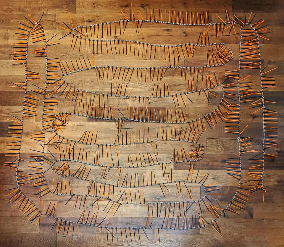A long silver-colored chain decorated with orange and black cable ties forms looping rows on a wooden floor.