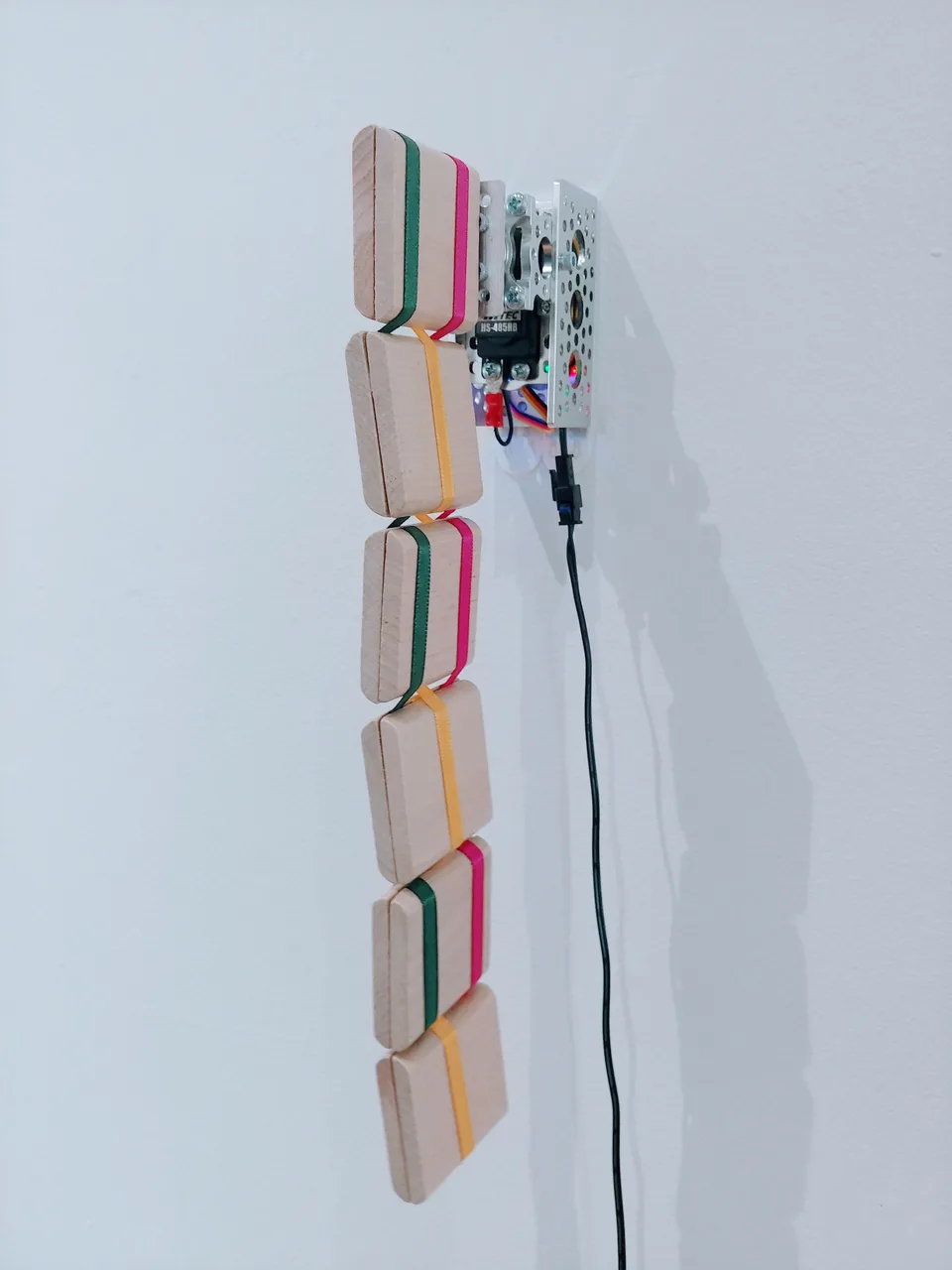 A chain of wooden blocks joined by colored ribbons hangs from a wall-mounted motorized mechanism.