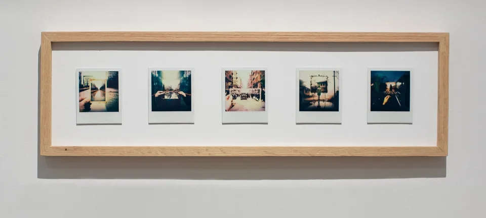 Five Polaroids in a long wooden frame show streets and buildings with hands holding smaller framed views inside the scenes.