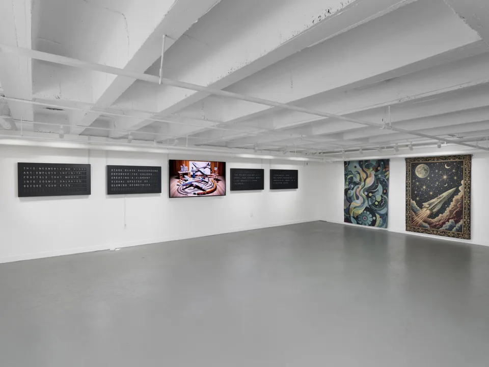 Four split-flap displays and a monitor share a corner of the white gallery with two colorful tapestries.