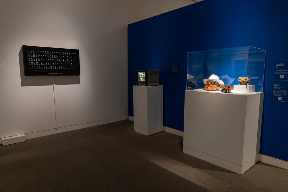 The split-flap criticism board hangs beside a monitor and a vitrine containing a cowboy puppet head and two small cardboard robots.