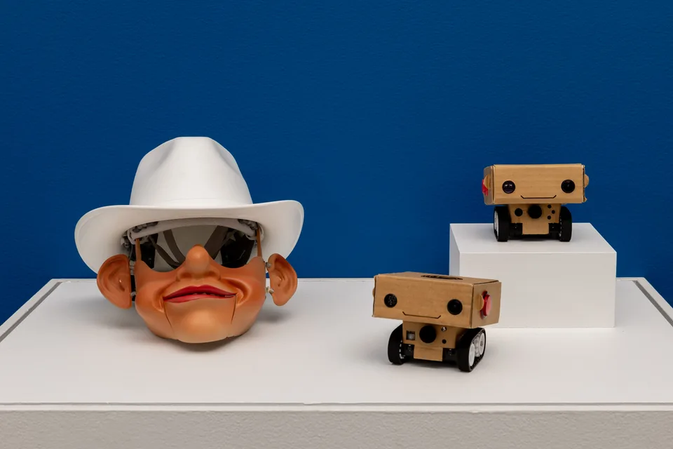 A robotic character head in a white cowboy hat displayed beside two small cardboard BlabDroids on white plinths.